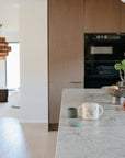 A modern kitchen with a marble countertop holds a white mug, green cup, and the mushie Snack Cup—a spill-proof silicone snack container for toddlers. In the background, white chairs and a copper chandelier shine in natural light.