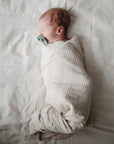 Newborn wrapped in mushie Organic Cotton Muslin Swaddle Blanket, lying with pacifier.
