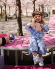 Young girl pours tea from Mushie's Silicone Tea Play Set at a pink table under blooming trees.