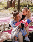 A smiling woman holds a baby with a FRIGG Daisy Natural Rubber Pacifier by pink benches.