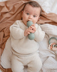 A baby in a cream knit outfit lies on a soft blanket, holding and chewing on a green food-grade silicone teething toy. A mushie Silicone Baby Rattle Toy rests nearby as the baby gazes up, captivated by the sensory experience.