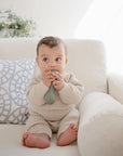 A baby in a beige knit outfit sits on a white couch, holding and chewing on the mushie Silicone Baby Rattle Toy. A decorative pillow and plant enhance the cozy, sensory atmosphere in the background.