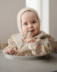 A baby sits in a high chair, smiling and holding a spoon, wearing mushie's Long Sleeve Bib along with a floral outfit and knitted bonnet.