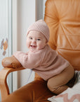 A smiling baby in a pink knit hat and sweater enjoys a mushie Suction Spinner Toy on a tan leather chair. A colorful cushion and window with flower decals appear in the background as the baby looks at the camera.