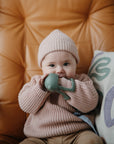 A baby in a pink knit sweater and hat smiles on a tan leather chair, holding the mushie No-Drop Cactus Teether (Dried Thyme). A colorful patterned pillow sits beside them.