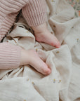 Close-up of a baby’s feet resting on the mushie Organic Cotton Muslin Swaddle Blanket, featuring small yellow and gray designs. The baby wears pink knitted pants, highlighting the blanket’s soft, patterned organic cotton fabric.
