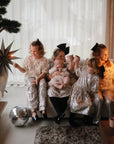 Five young children in glittery outfits sit on a sofa, holding sparklers, a baby, and colorful mushie Dinnerware Cups. A disco ball sparkles on the floor by large windows with sheer curtains, adding to the festive mood.