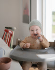 A smiling toddler in a gray knit beanie and beige bib sits in a high chair eating with mushie Silicone Toddler Starter Spoons. Food is scattered on the tray and bib. Sunlight streams in through a nearby open door, brightening the room.