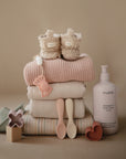 A stack of folded baby clothes topped with fuzzy booties, surrounded by a pink shampoo bottle, mushie Silicone Toddler Starter Spoons for self-feeding, teething toys, and a pacifier, all on a neutral background.