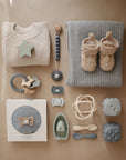 Flat lay of baby items on a tan background, including a folded cream sweater, blue blanket, fuzzy booties, toys, pacifier clip, silicone teether, mushie Silicone Toddler Starter Spoons for independent feeding, and a boxed toy set in neutral and blue tones.