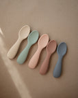 Mushie Silicone Toddler Starter Spoons—five pastel spoons in beige, green, pink, peach, and gray—are arranged on a light brown surface with natural light, perfect for encouraging independent feeding.