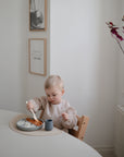A young child eats rice and sauce with a spoon from the mushie Silicone Suction Plate at a bright, minimally decorated table. A small cup sits beside the plate, and there’s framed art and a vase with red leaves nearby.