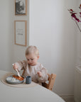 A young child with short blond hair sits at a light-colored table, eating from a plate using the mushie Dinnerware Fork and Spoon Set. A small cup sits on a placemat, with minimalist decor and a plant in the background.