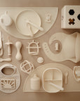A top-down view displays cream-colored baby feeding items and toys, including mushie Silicone Toddler Starter Spoons, plates, bottles, a bib, a sectioned tray, a shape-sorting cube, and silicone baby spoons on a beige surface.