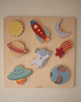 A wooden puzzle featuring colorful pieces: the sun, a rocket, the moon, Saturn, an astronaut, a comet, a flying saucer, and planet Earth, all arranged on a square wooden board.