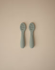 Two light green Mushie First Feeding Baby Spoons with textured silicone tips and embossed handles are placed side by side on a neutral beige background.