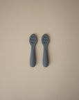 Two gray Mushie First Feeding Baby Spoons with textured teething tips are placed parallel on a beige background, with the Mushie brand name embossed on their handles.