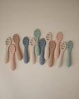 Twelve pastel Mushie First Feeding Baby Spoons—pink, blue, green, and cream—are neatly arranged. Each features a flat, slotted head and rounded handle for easy self-feeding.