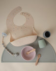 The mushie Silicone Placemat is arranged with a speckled bib, two small cups, a pink bowl and spoon, and a pacifier clip—an ideal choice for stylish baby mealtime essentials.