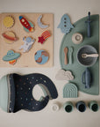 A flat lay of space-themed baby items, including a planet and rocket wooden puzzle, mushie Rocket Rattle Teether, silicone bib with space prints, silicone bowls and utensils, plus small pastel-colored cups.