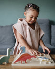 A young girl with braided hair and pink clips, wrapped in a mushie Bear Poncho Towel made of soft organic cotton, plays with wooden dinosaur toys on a table in a cozy living room with a gray sofa in the background.