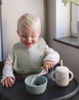 A smiling toddler with blonde hair sits in a high chair, using the mushie Dinnerware Cutlery Set's non-toxic spoon to eat from a bowl, with a sippy cup and a light green bib with blue penguins nearby by the window.