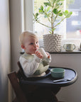 A young blond child sits in a high chair by a window, wearing a bib and holding the mushie Bluey x Mushie Snack Cup. A green bowl and spoon are on the tray with other baby feeding essentials, and a potted plant is on the windowsill behind.