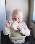 A young blonde child in a high chair smiles while sipping from the Mushie Bluey x Mushie Silicone Training Cup + Straw, a food grade silicone cup with an animal design, enjoying natural light streaming through a nearby window.