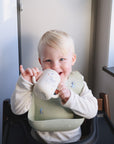 A young blonde child in a high chair smiles while sipping from the Mushie Bluey x Mushie Silicone Training Cup + Straw, a food grade silicone cup with an animal design, enjoying natural light streaming through a nearby window.