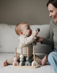 A baby smiles at a woman while holding a toy on the couch. In front, the Mushie Wooden Multi Shape Stacker with pastel rings boosts joy and helps develop fine motor skills during playtime.
