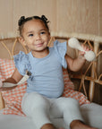 A young child with curly hair smiles on a wicker chair, enjoying a fun sensory experience while holding the mushie Silicone Baby Rattle Toy in one hand and a pacifier clip in the other.