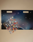 An open space-themed folder with starry sky artwork rests on a beige surface, revealing mushie Reusable Sticker Set—colorful puffy rockets, planets, and spaceships, perfect for sticker books or creative play.