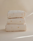 Three mushie Extra Soft Muslin Crib Sheets, cream-colored, stacked on a neutral sun-patterned background.
