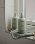 Two green pump bottles labeled mushie baby body lotion and shampoo & body wash sit on a bathroom windowsill with a green washcloth, a pacifier, and a cream-colored sponge nearby.