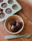 Three berry cubes in a Mushie Baby Food Freezer Tray with baby spoon on wood.