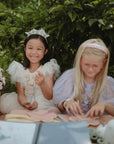 Two girls in princess dresses craft outdoors with a mushie Silicone Sensory Toy, one smiles at camera.