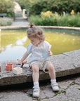 A young child plays by a pond with the mushie Mix and Match Present Toy, colorful wooden blocks designed to encourage fine motor skills, surrounded by lush greenery and blooming flowers on a sunny day.