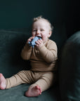 A smiling baby in beige pajamas sits on a green sofa, holding and chewing on the mushie Rocket Rattle Teether in blue while looking at the camera.
