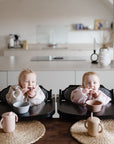 Two toddlers sit side by side at a dining table, each wearing a mushie Long Sleeve Bib. Their snacks, bowls, and cups are in front of them. A modern minimalistic kitchen is visible in the background.