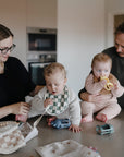 A woman and man care for two babies in a kitchen. One baby, wearing a mushie Chunky Knit Sweater, plays with toys on the counter, while the man holds another baby with a teething ring. Items are spread on the countertop.