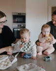 A woman and man care for two babies in a kitchen. One baby, wearing a mushie Chunky Knit Sweater, plays with toys on the counter, while the man holds another baby with a teething ring. Items are spread on the countertop.