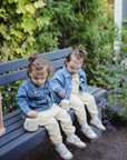 Two toddlers with brown hair in ponytails, wearing denim jackets and yellow pants, sit on a garden bench holding spill-proof mugs. A mushie Snack Cup and an abacus are beside them, surrounded by lush greenery.