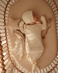 A baby sleeps peacefully in a beige bassinet, wrapped in the mushie Star Lovey Blanket, surrounded by soft bedding, a star-shaped pillow, and a wooden rattle. Warm sunlight softly illuminates the scene.