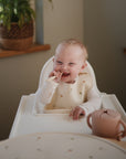 A smiling baby in a high chair wears a patterned bib and light long-sleeve shirt, holding one hand by their mouth. On the tray is a pink sippy cup and a mushie Silicone Placemat made from food-grade silicone. A window and potted plant are behind.