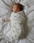 A newborn baby is swaddled in a mushie Organic Cotton Muslin Swaddle Blanket with whale prints, lying on a white bed, with an organic cotton pacifier placed nearby to the left.