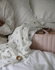 A person sits on a bed with a white comforter beside a soft white blanket featuring small blue designs and a light pink mushie Diaper Caddy filled with baby items.