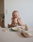 A smiling young child with blonde pigtails sits at a table, holding a pink cup. Snacks rest on the mushie Silicone Placemat, made from food-grade silicone, offering style and safety in a bright, minimal space.