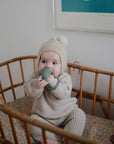 A baby in a beige knit outfit and hat sits in a wooden crib with a patterned mattress, holding and chewing on the mushie No-Drop Cactus Teether in Dried Thyme. A framed picture hangs on the wall behind.