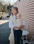 A woman smiles outdoors by a pink wall, holding a baby in a beige wrap. The alert baby uses a FRIGG Natural Rubber pacifier from the FRIGG 6-Pack while sitting near white chairs and a table on this sunny day.