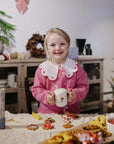 Smiling child holds Mushie Silicone Training Cup + Straw at a table with cookies, holiday decor.